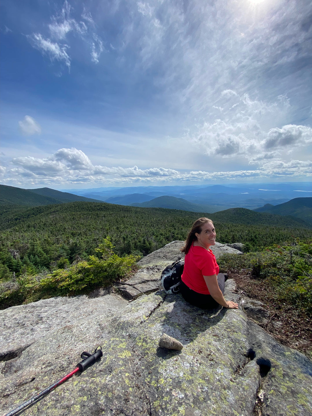 Person sitting on a rocky outcrop in the Adirondacks with a scenic mountain view and blue sky.