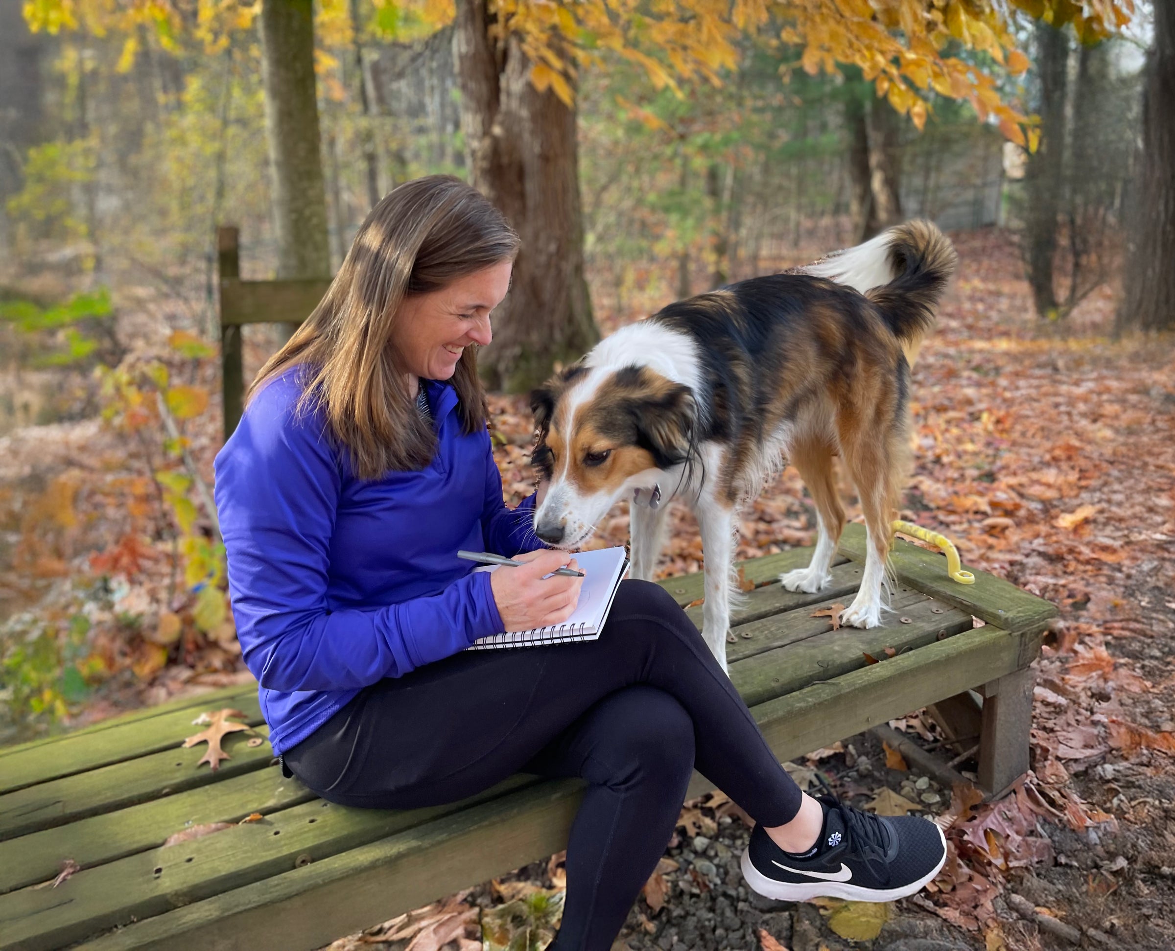 Woman sitting on a wooden bench with a dog, writing in a notebook in a forest setting.